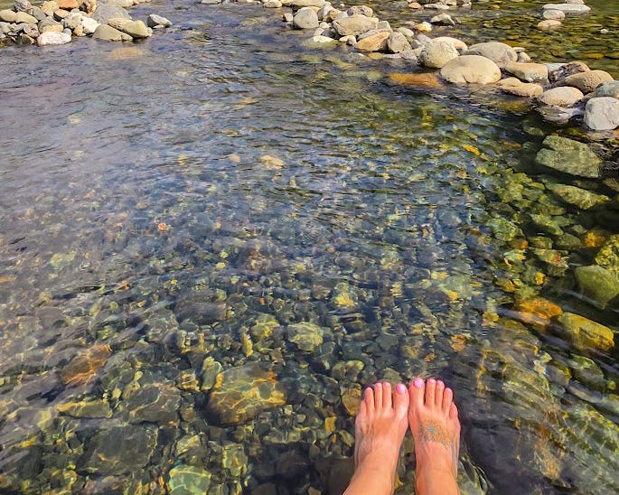 Feet dangling in a clear river lined with round stones.